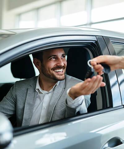 Modulo texto Comprar un coche - Chico joven en su coche sentado mientras le dan las llaves de su coche nuevo